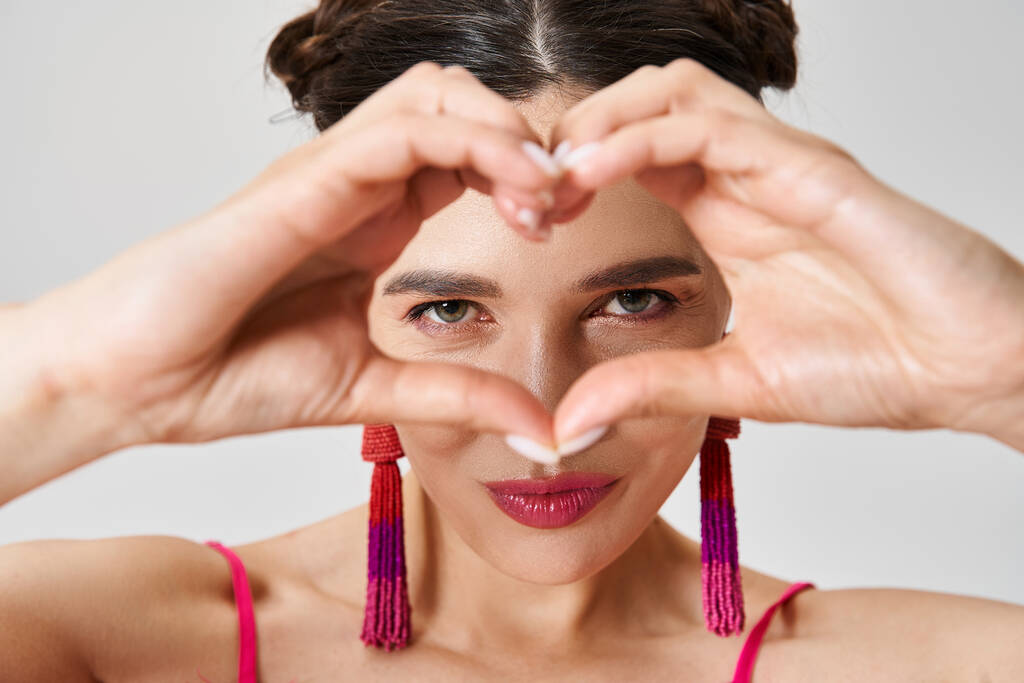 Joyful brunette woman wearing pink feather dress showing a hand heart and looking through it - Photo, Image