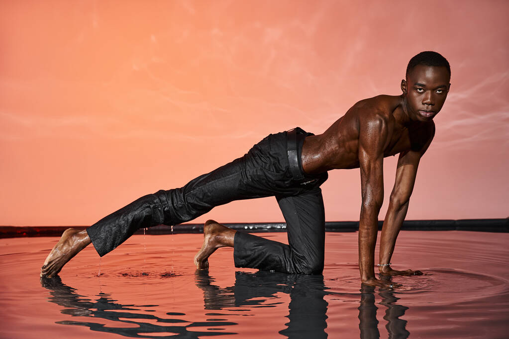 atraente sem camisa afro americano homem em preto molhado calças posando na água e olhando para a câmera - Foto, Imagem