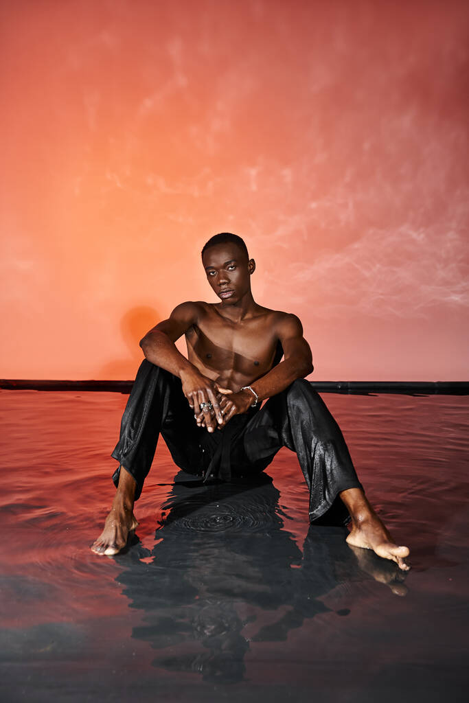 handsome african american man sitting topless on water surface and looking at camera, red lights - Photo, Image