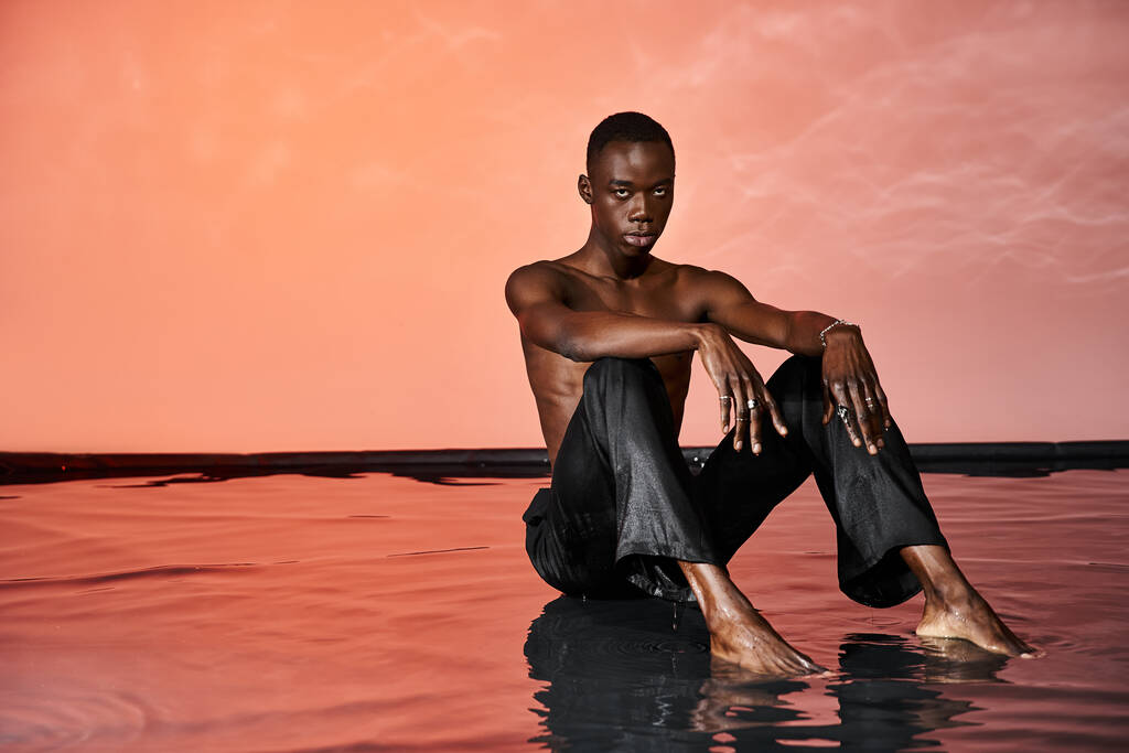 sedutor homem americano africano em calças molhadas sentado na superfície da água e olhando para a câmera, luz vermelha - Foto, Imagem