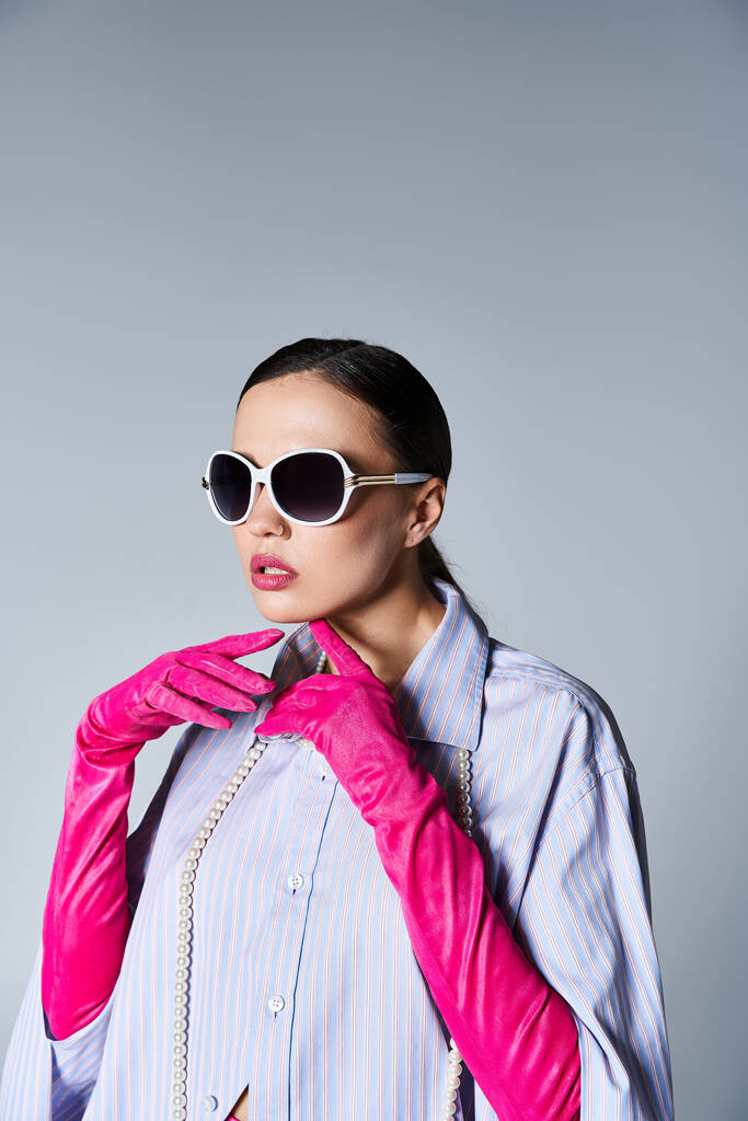 Όμορφη μελαχρινή γυναίκα σε μοντέρνα ενδυμασία και γυαλιά ηλίου αγγίζοντας το πηγούνι της στο στούντιο - Φωτογραφία, εικόνα