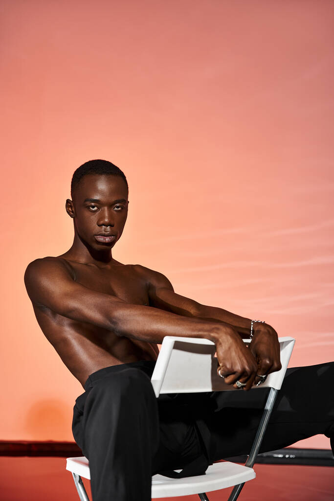 bonito sexy afro americano homem sentado sem camisa na cadeira e olhando para a câmera, luzes vermelhas - Foto, Imagem