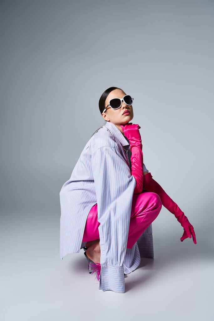 Με αυτοπεποίθηση μελαχρινή σε ροζ δερμάτινο παντελόνι και chic γυαλιά ηλίου κάθεται κομψά σε γκρι φόντο - Φωτογραφία, εικόνα