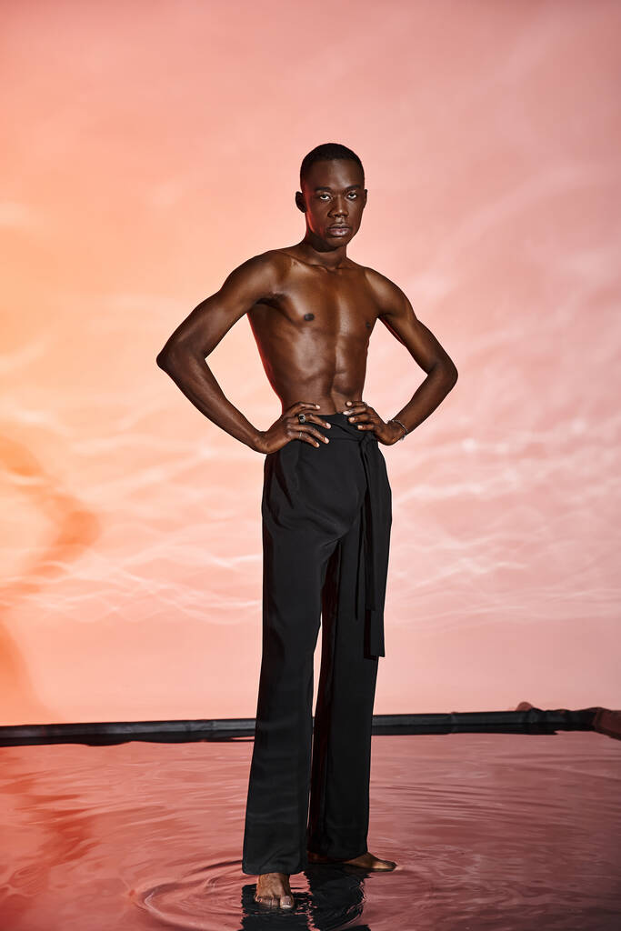 заманчивый африканский мужчина без рубашки стоит и смотрит в камеру в окружении красных фонарей - Фото, изображение