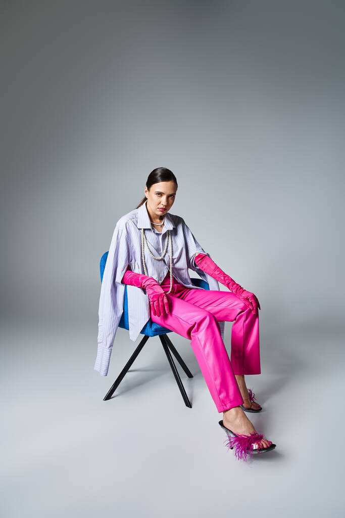 Шикарна брюнетка в стильному рожевому вбранні, торкаючись її ніг, сидить на стільці на сірому фоні - Фото, зображення