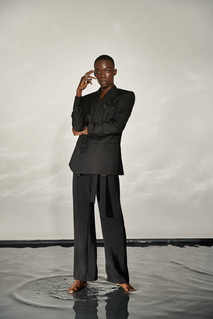 привабливий афроамериканський чоловік в чорному сексуальному костюмі позує привабливо на сірому водному фоні - Фото, зображення