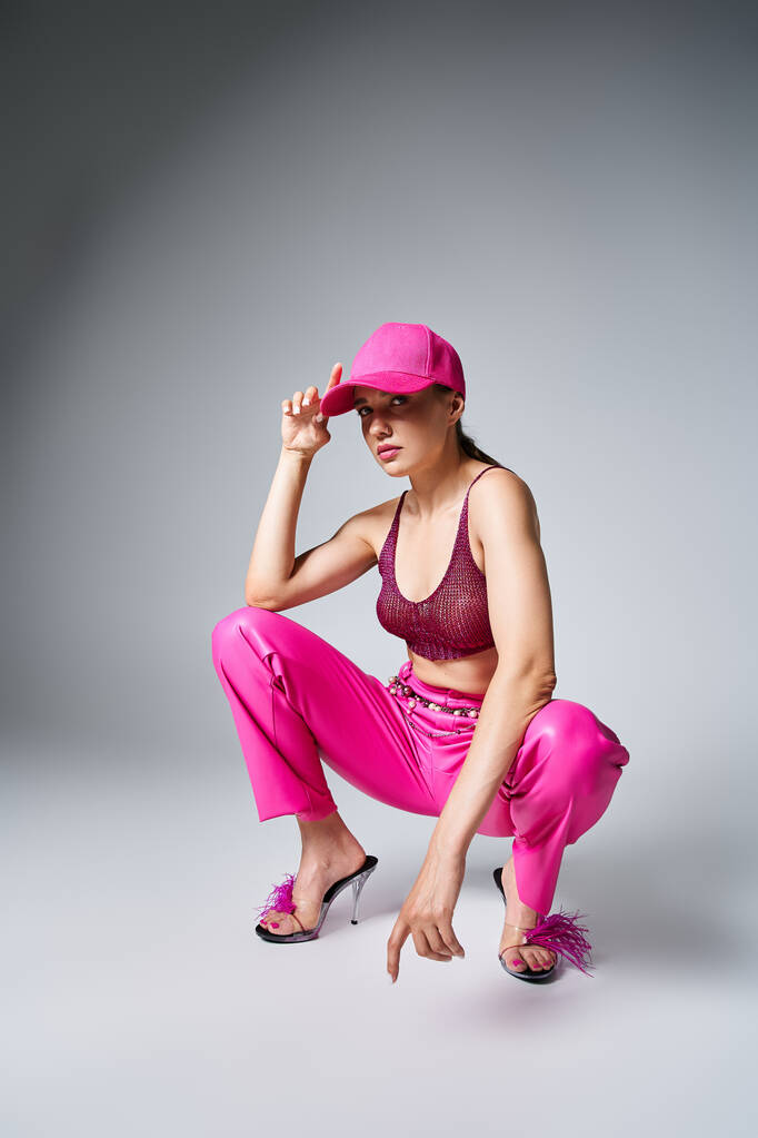 Fancy brunette woman in red top and pink trousers sitting and touching pink cap with one hand - Photo, Image