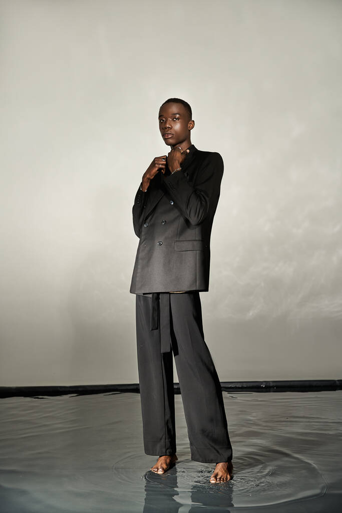 schöner afrikanisch-amerikanischer Mann im schwarzen sexy Anzug posiert attraktiv auf grauem wässrigem Hintergrund - Foto, Bild