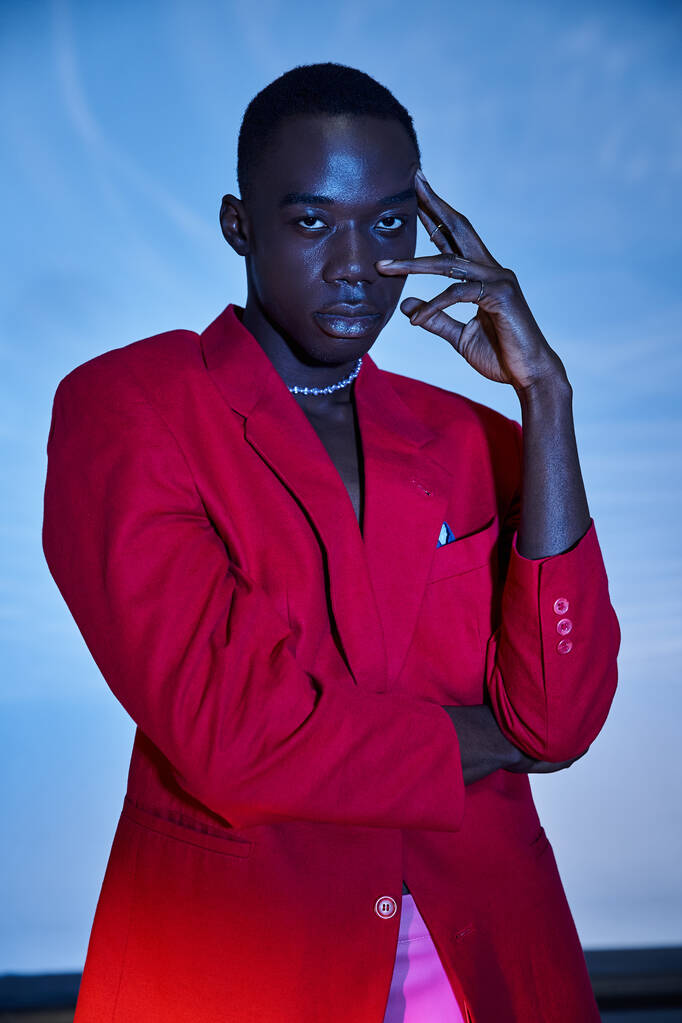 Kırmızı renkli ceketli yakışıklı Afro-Amerikalı adam mavi sulu arka planda kameraya bakıyor. - Fotoğraf, Görsel
