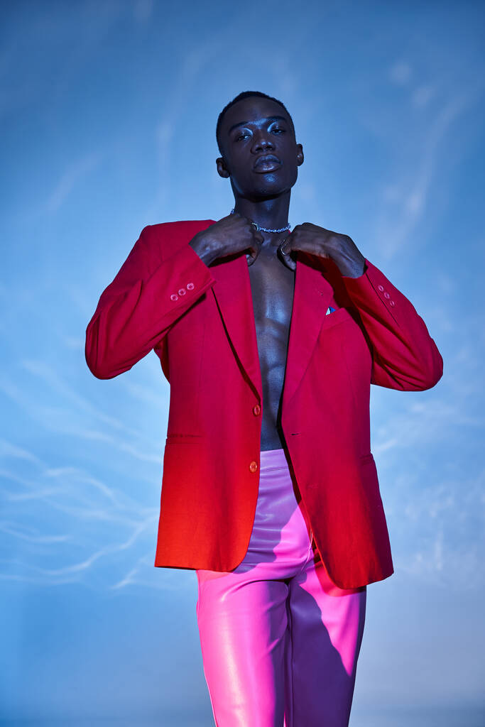 tentador hombre afroamericano con estilo en pantalones rosas y chaqueta roja posando sobre fondo azul acuoso - Foto, Imagen