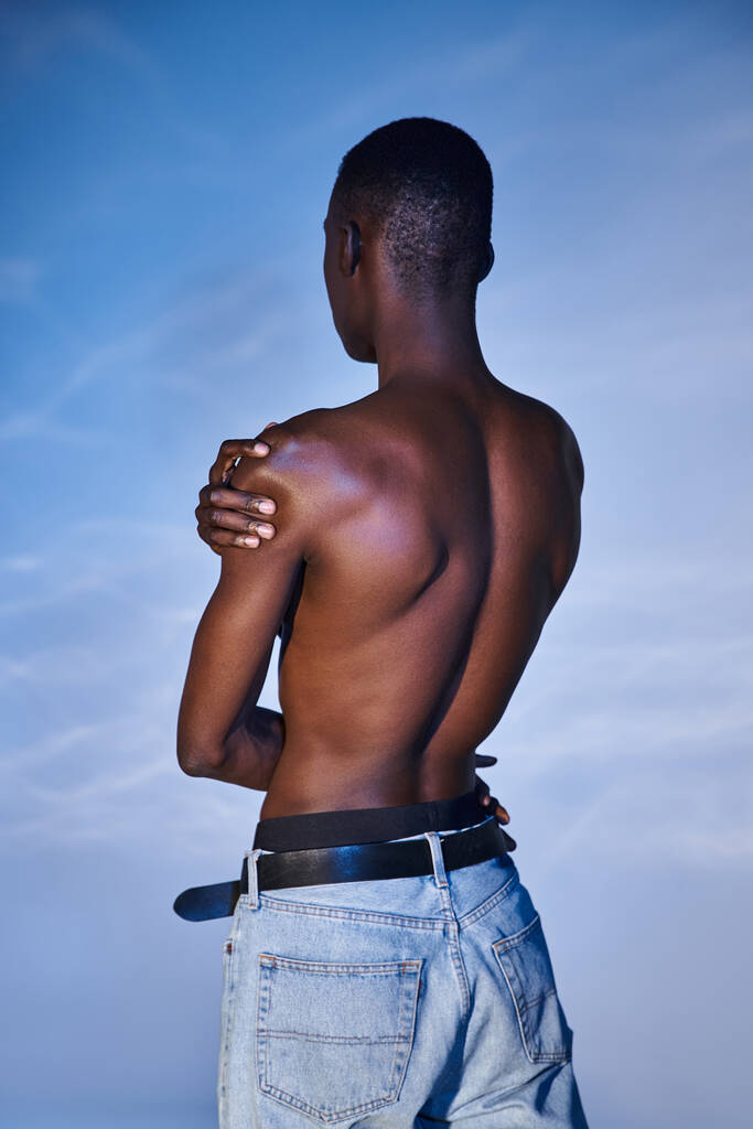 widok z tyłu modny Afrykański człowiek bez koszuli w stylowych dżinsach na niebieskim tle wodnistej - Zdjęcie, obraz