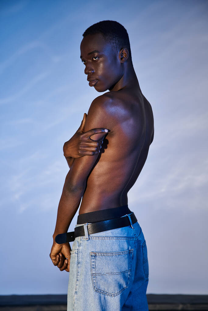 Африканский мужчина без рубашки в стильных джинсах позирует на синем водянистом фоне и смотрит в сторону - Фото, изображение