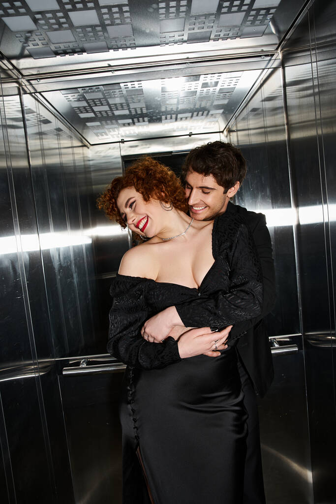 エレベーターで抱擁エレガントな黒の服装で楽しいボーイフレンドとガールフレンドを魅了, セクシーなカップル - 写真・画像