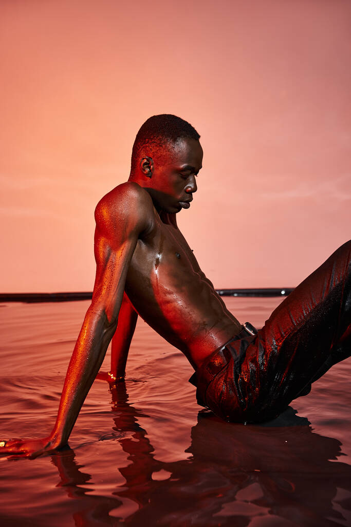 sexy uomo africano americano senza maglietta in pantaloni neri bagnati seduto sulla superficie dell'acqua a luci rosse - Foto, immagini