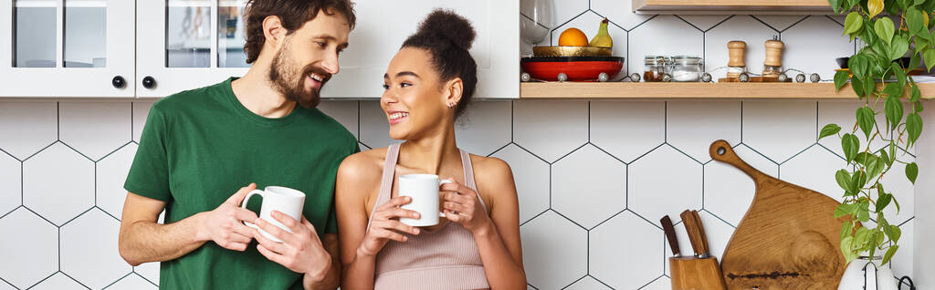 atraktivní veselý rozmanitý pár v homewear těší horké kávy a při pohledu na sebe, banner - Fotografie, Obrázek