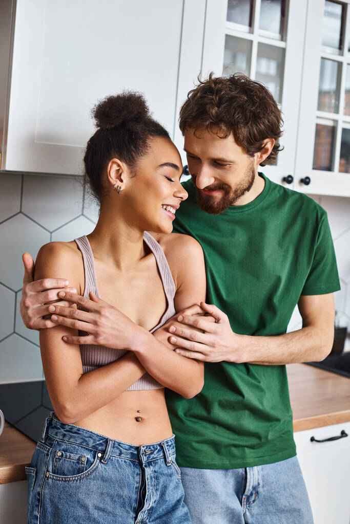 cheerful good looking diverse couple in homewear hugging each other lovingly in kitchen at home - Photo, Image