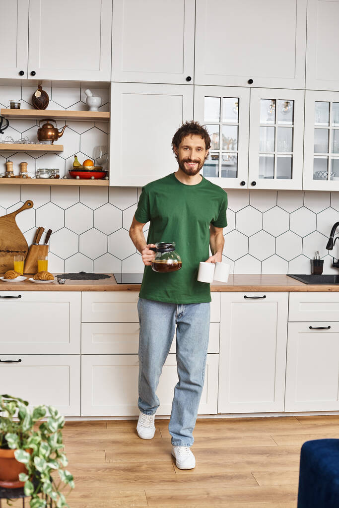 knappe vrolijke man in casual homewear houden koffie en glimlachen op camera terwijl in de keuken - Foto, afbeelding
