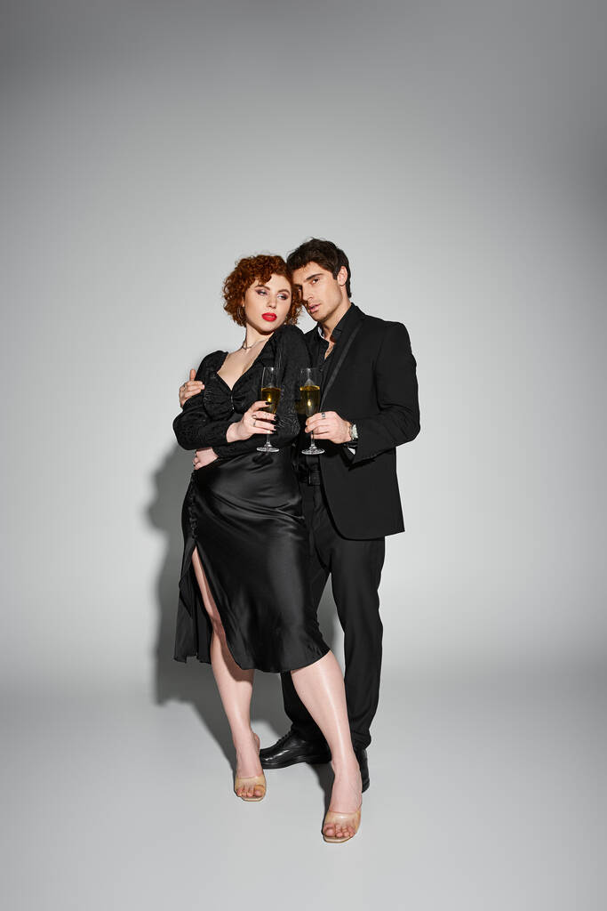 piękna kobieta w czarnej sukience przytula swojego przystojnego chłopaka na szarym tle, szampan w rękach - Zdjęcie, obraz