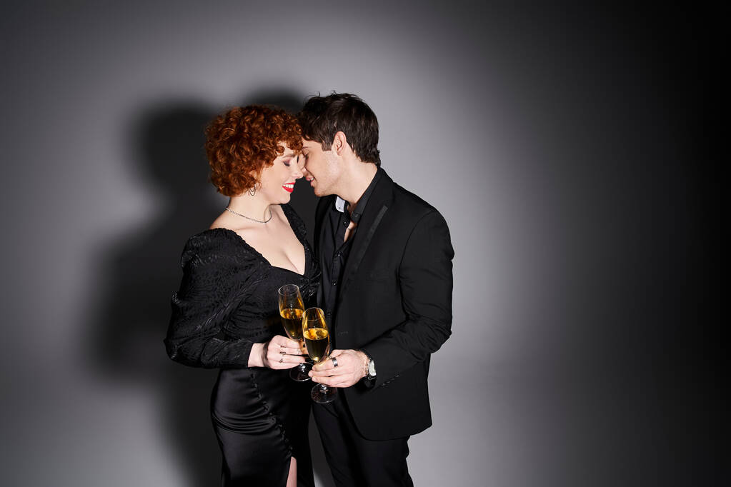привлекательная женщина в черном платье обнимает своего красивого парня на сером фоне, шампанское в руках - Фото, изображение