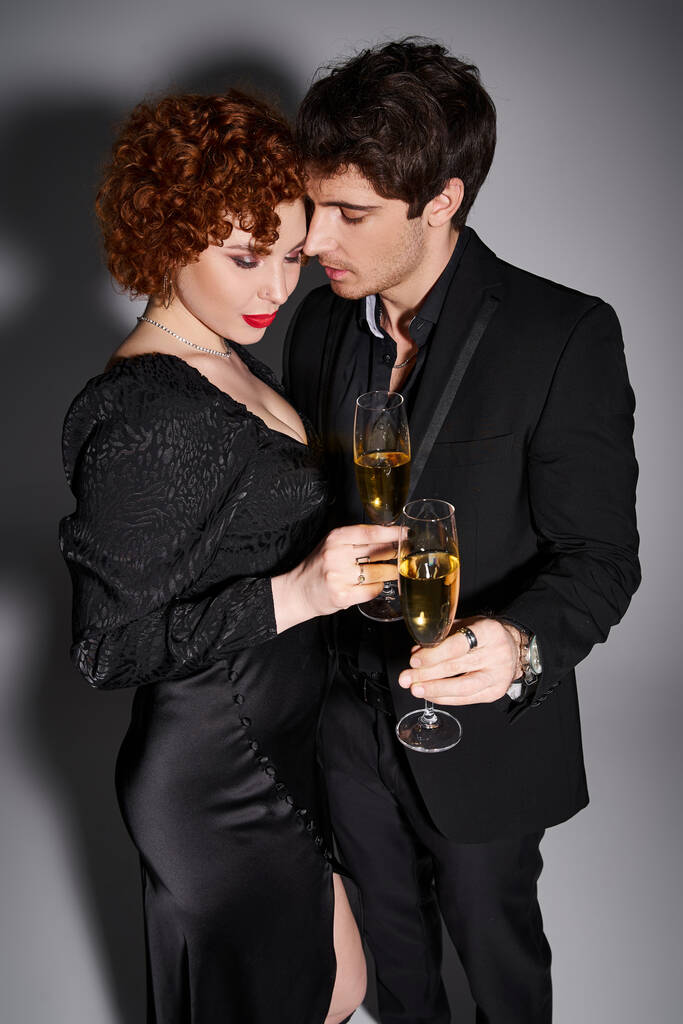 charming woman in black dress hugging her handsome boyfriend on gray backdrop, champagne in hands - Photo, Image