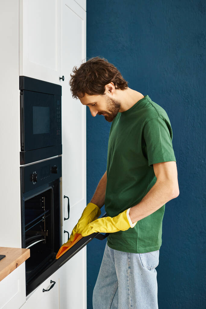 handsome cheerful man in dark green t shirt and gloves cleaning modern oven with yellow rag - Photo, Image