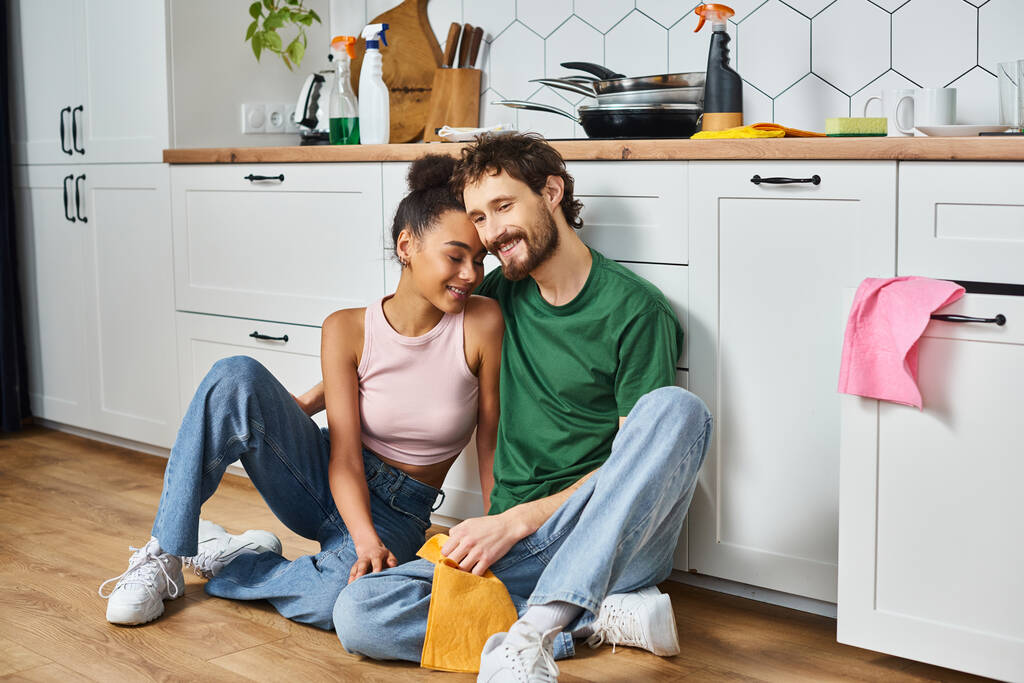 interracial veselý pár v ležérní oblečení sedí na podlaze při čištění a šťastně se usmívá - Fotografie, Obrázek