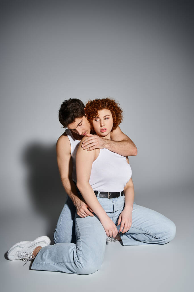 kuszące mężczyzna i kobieta w niebieski dżinsy siedzi na podłodze razem i przytula ciepło, sexy para - Zdjęcie, obraz