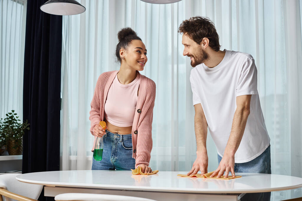 allegro attraente coppia diversificata in comodi casalinghi sorridenti a vicenda durante la pulizia a casa - Foto, immagini