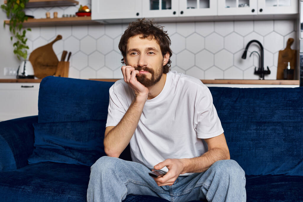 hombre guapo aburrido en acogedora ropa de casa sentado en el sofá con control remoto en la mano y viendo la televisión - Foto, imagen