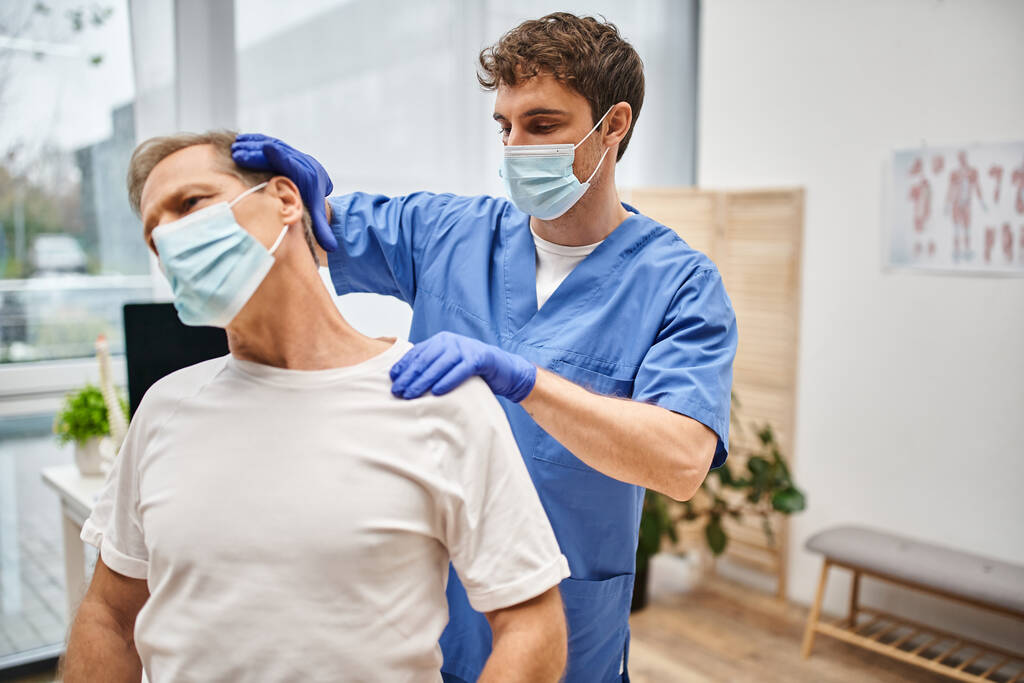 gewijd goed uitziende arts met masker stretching zijn patiënt tijdens een afspraak in het ziekenhuis afdeling - Foto, afbeelding