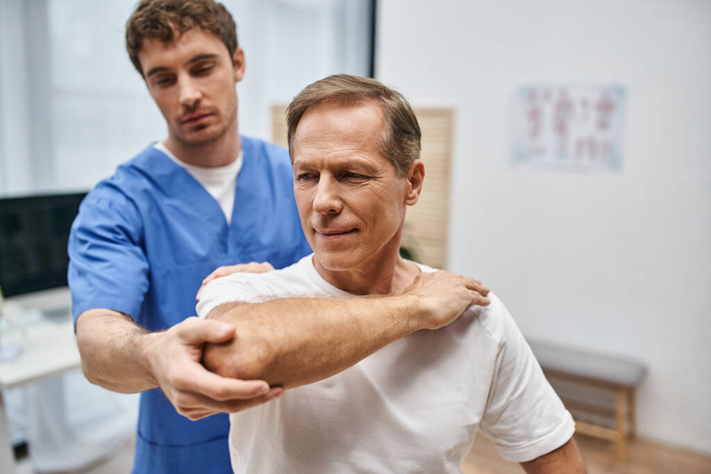 зрілий чоловік в повсякденному вбранні розтягує м'язи за допомогою свого відданого лікаря, реабілітація - Фото, зображення