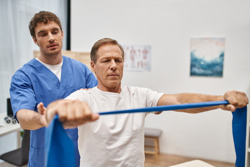 jóképű orvos kék egyenruhában segít az érett beteg ellenállás szalag rehabilitáció - Fotó, kép