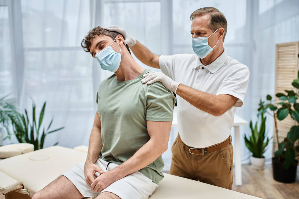 beau réhabilitologue mature avec masque et gants massant le cou de son patient à l'hôpital - Photo, image