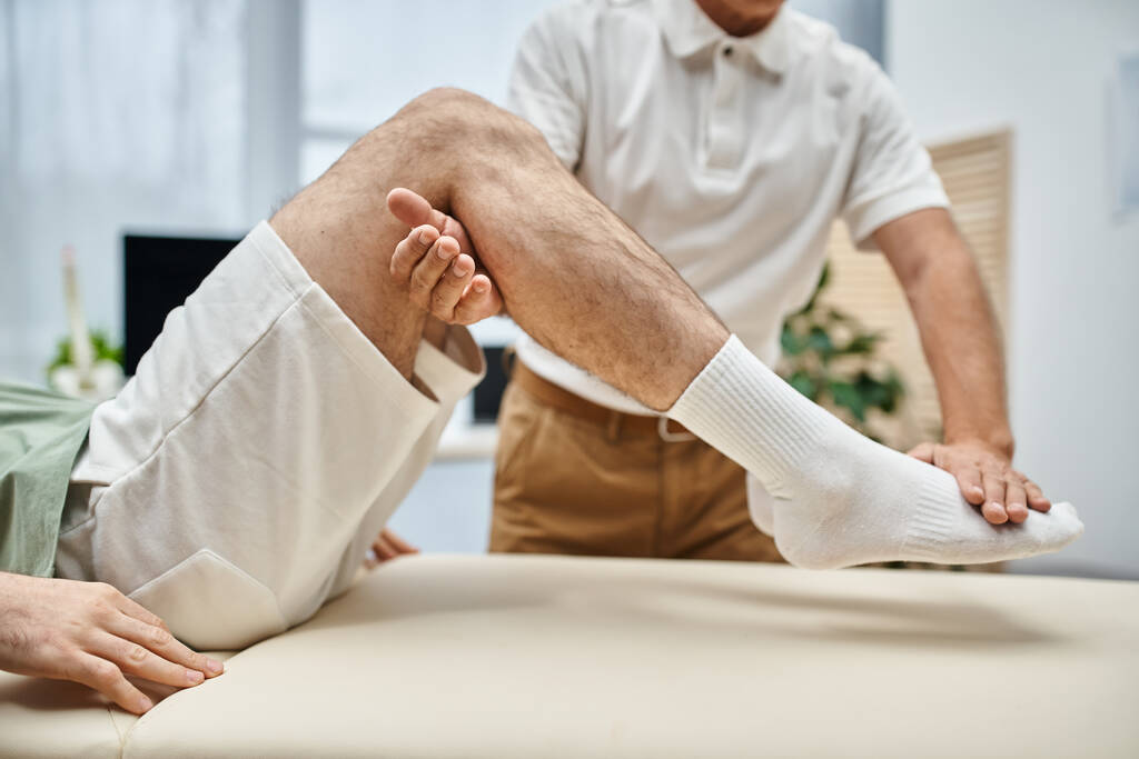 goed uitziende volwassen arts controleren van knieën van zijn patiënt in het ziekenhuis, revalidatie concept - Foto, afbeelding