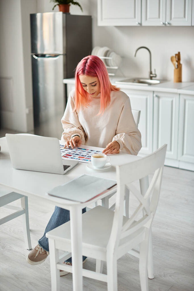 vrolijke jonge vrouw in casual kleding met roze haar werken met laptop en papierwerk met beelden - Foto, afbeelding