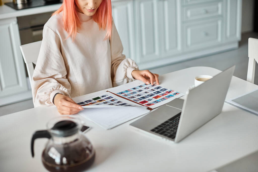 Ausgeschnittene Ansicht einer Frau in Freizeitkleidung, die neben ihrem Laptop und Kaffee Papiere mit Bildern hält - Foto, Bild