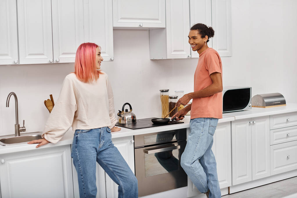 bonito alegre afro-americano homem cozinhar um pouco de comida e sorrindo para sua namorada de cabelos cor de rosa - Foto, Imagem