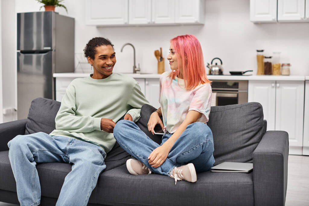 aantrekkelijk vrolijke interraciale paar in homewear zitten op de bank en glimlachen gelukkig naar elkaar - Foto, afbeelding