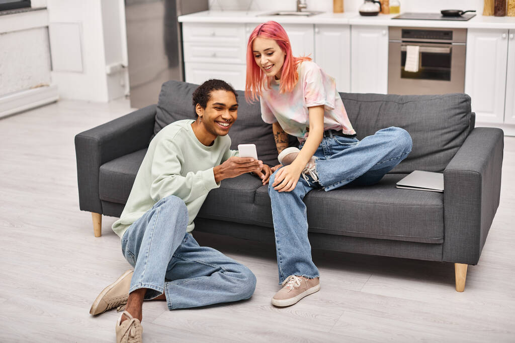 fröhlich ansprechendes gemischtrassiges Paar in Homewear, das im Wohnzimmer sitzt und aufs Smartphone schaut - Foto, Bild