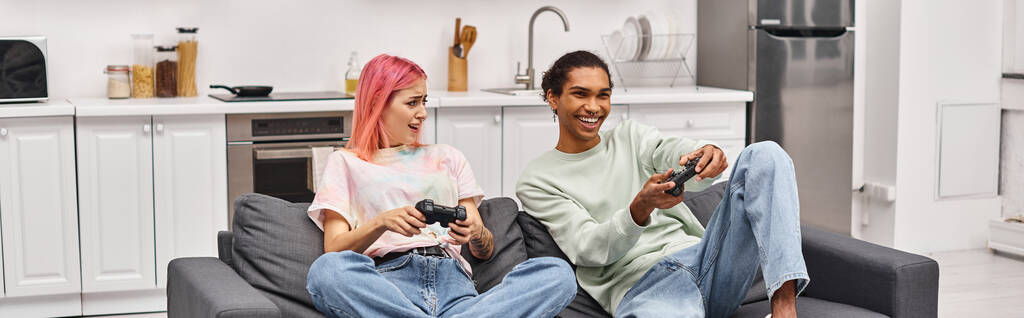 joyeux couple multiracial dans des vêtements confortables assis et en utilisant des manettes de jeu pour jouer à des jeux dans le salon - Photo, image