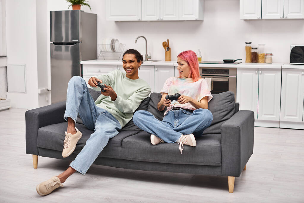 Веселая мультикультурная пара в уютной домашней одежде сидит и играет в видеоигры с джойстиками дома - Фото, изображение