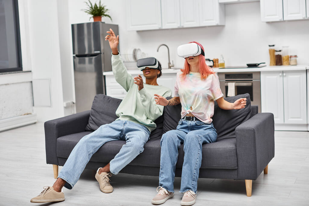 přitažlivý mladý mezirasový pár v pohodlném oblečení sedí na pohovce doma ve svých VR sluchátkách - Fotografie, Obrázek