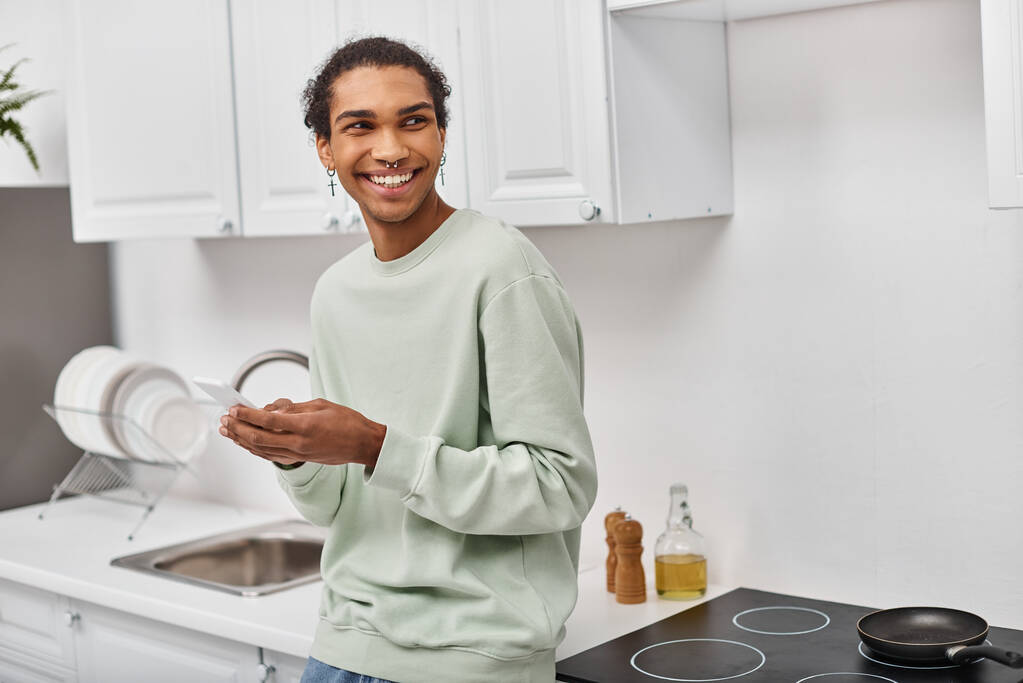 guapo hombre afroamericano alegre en suéter casual blanco sosteniendo teléfono inteligente y mirando hacia otro lado - Foto, Imagen
