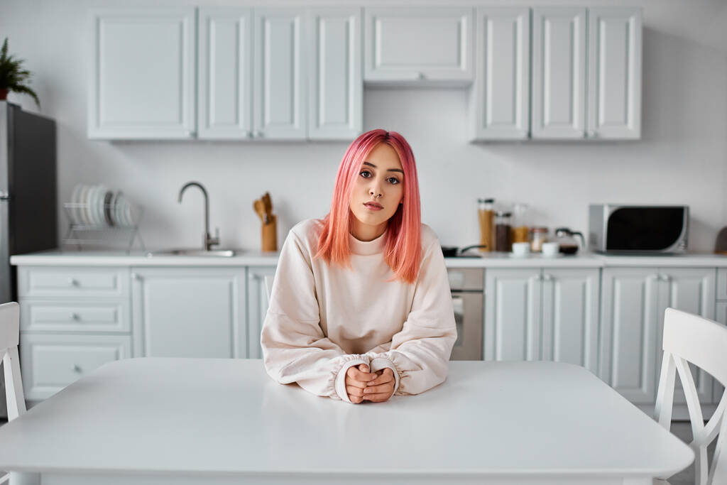 テーブルに座ってカメラを見ている居心地の良い家のウェアのピンクの髪の魅力的な若い女性 - 写真・画像
