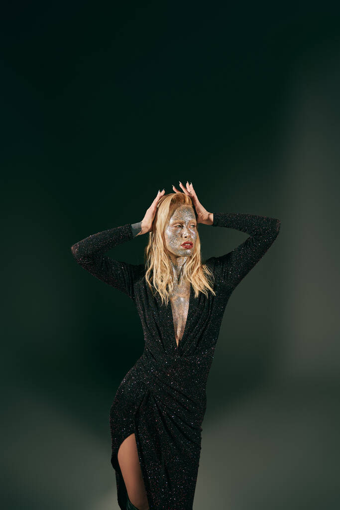 блондинка з блиском на обличчі позує в блискучій сукні з руками біля волосся на чорному тлі - Фото, зображення