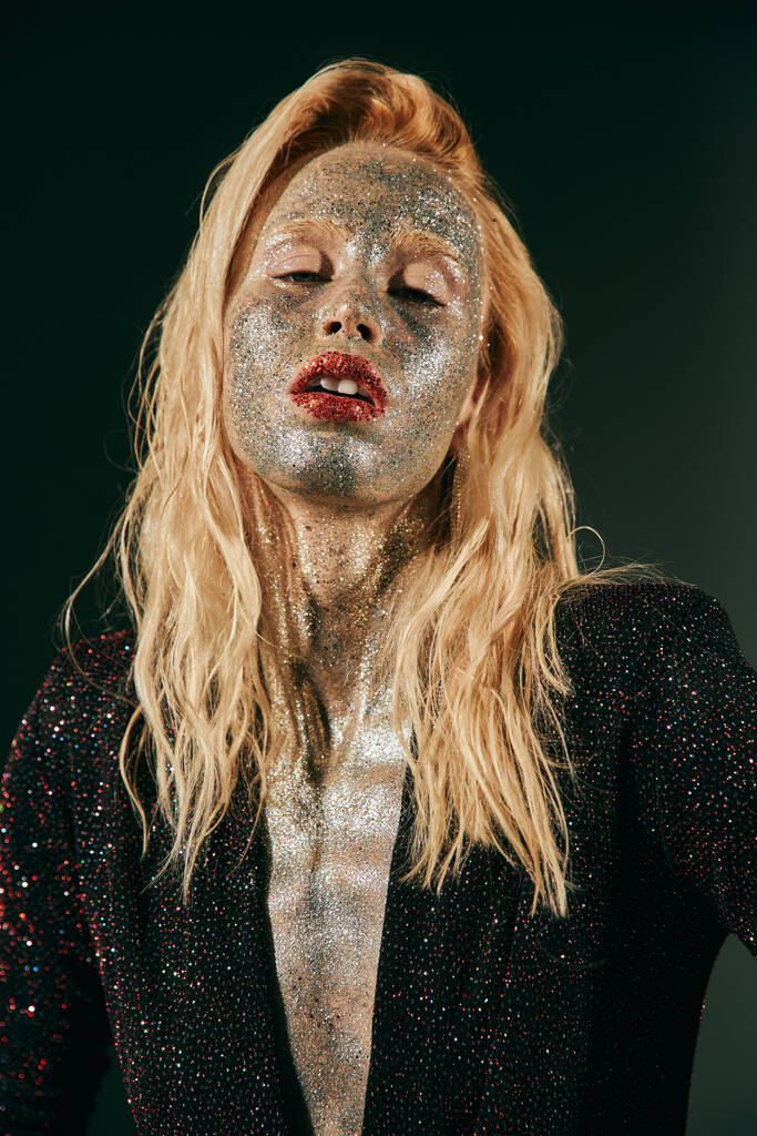 πορτρέτο της ξανθιάς γυναίκας με glitter στο πρόσωπο και το σώμα ποζάρουν σε γυαλιστερό φόρεμα σε μαύρο φόντο - Φωτογραφία, εικόνα