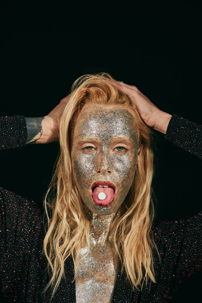 προκλητική νεαρή ξανθιά γυναίκα με glitter στο πρόσωπο δείχνει γλώσσα με χάπι σε μαύρο φόντο - Φωτογραφία, εικόνα