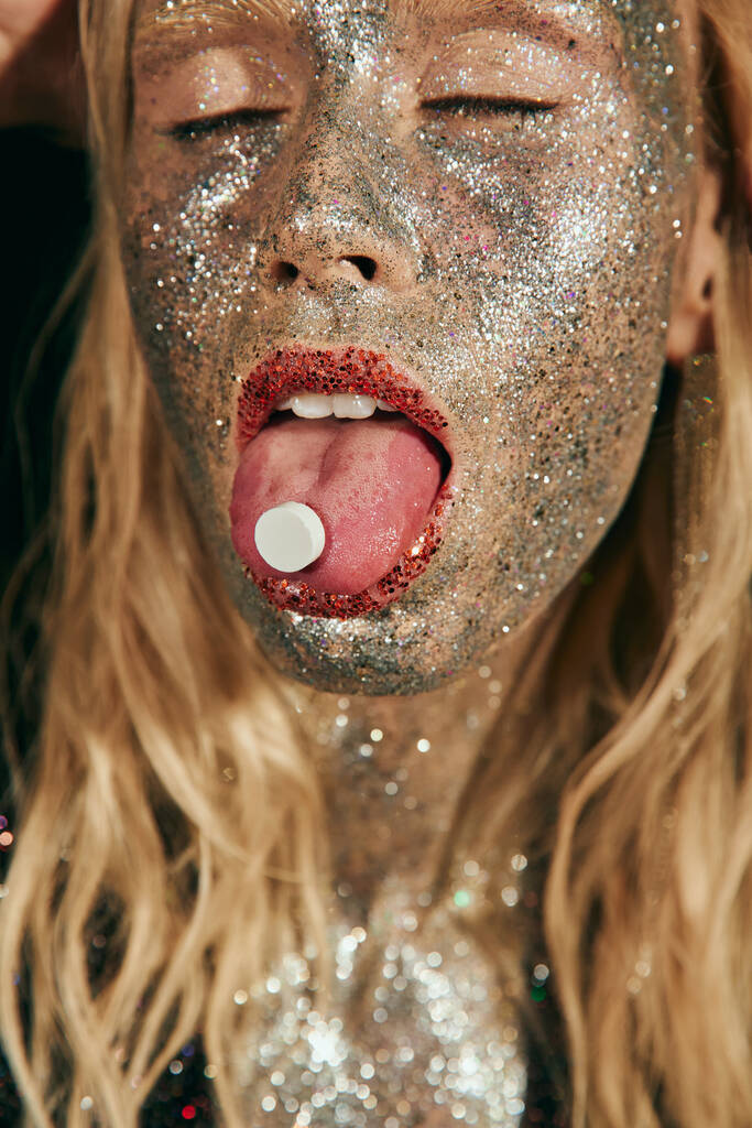 крупним планом молода жінка з блискучим макіяжем і закритими очима, показуючи язик з таблеткою на чорному - Фото, зображення