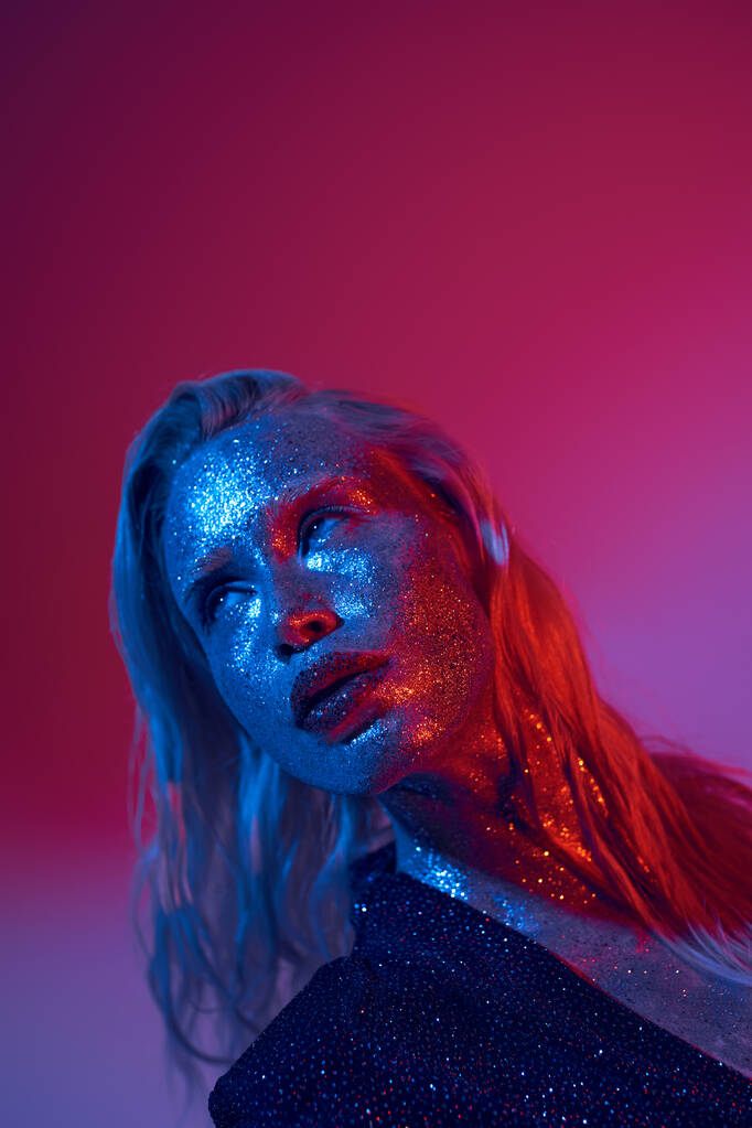 blonde and shiny woman with glitter on face and sparkling outfit posing on vibrant neon background - Photo, Image