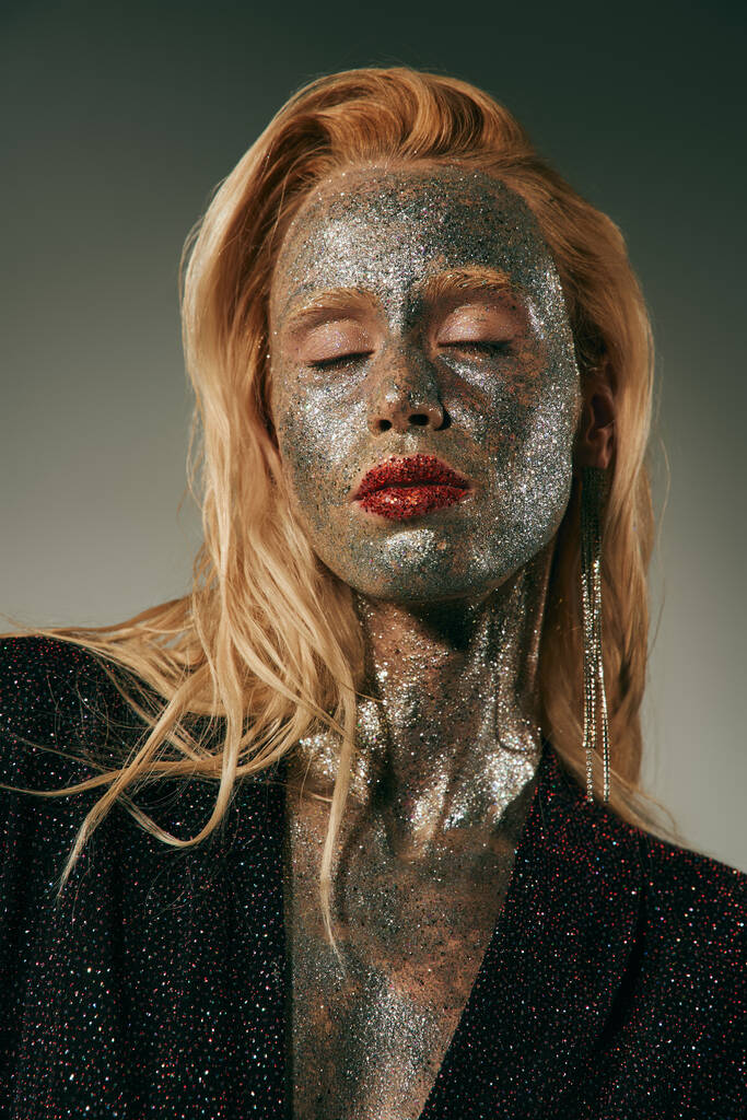 alluring young woman with closed eyes and glitter all over body and face posing on grey backdrop - Photo, Image